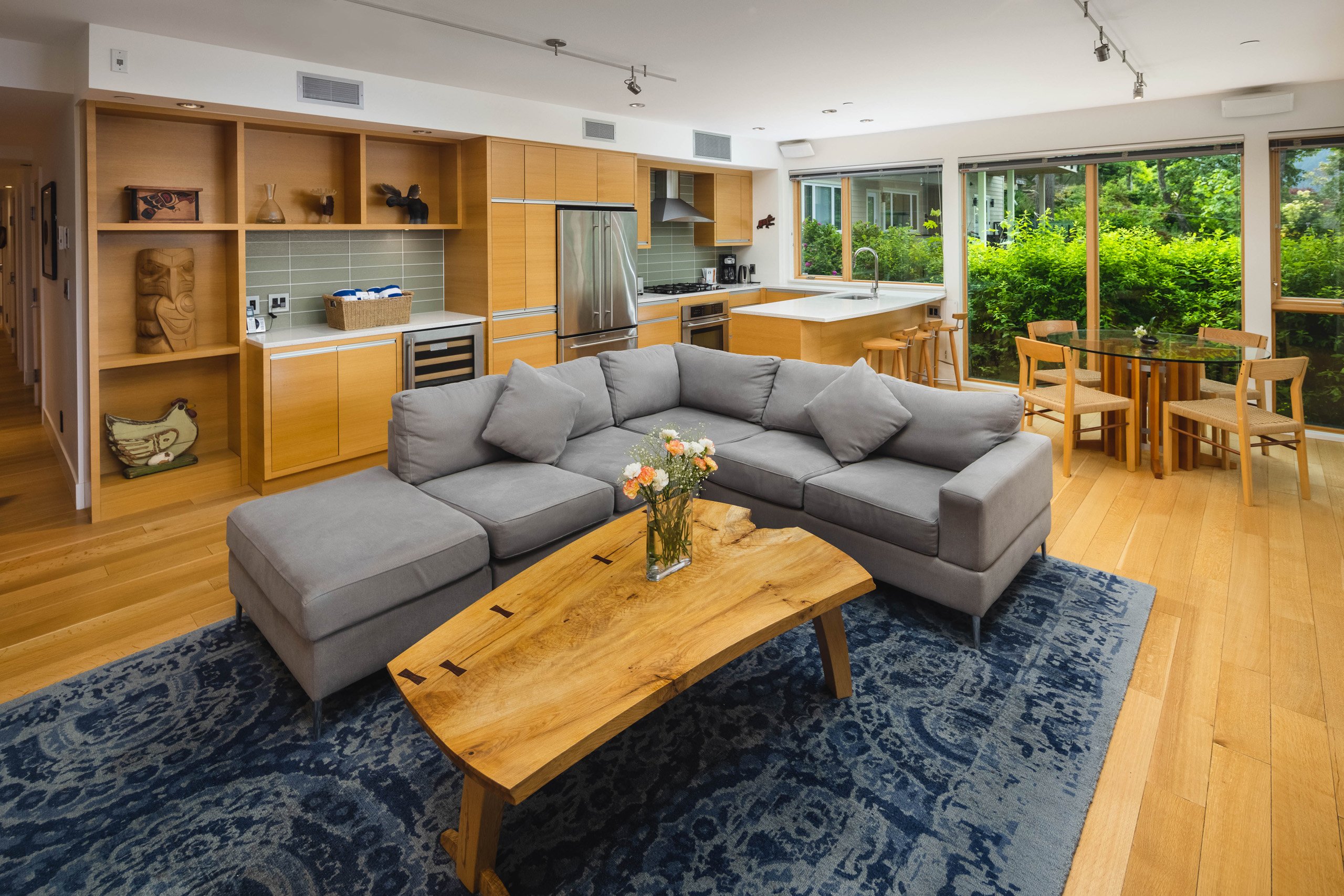 A modern open-plan living room and kitchen in this stylish villa features a gray sectional sofa, wooden coffee table, and blue rug. The kitchen boasts wooden cabinets and stainless steel appliances. Large windows reveal lush greenery alongside a wooden dining table.